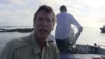 Clay Bryce talking to camera sitting in a small boat with man standing behind him steering  