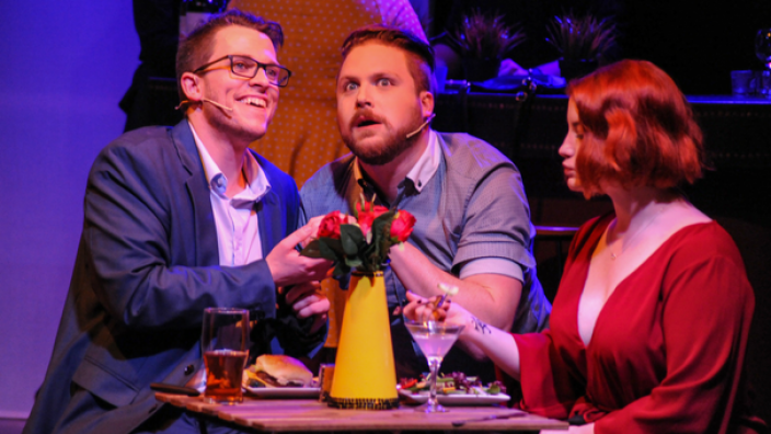Caption: First Date the Musical "Two men and a woman gathered around a table."