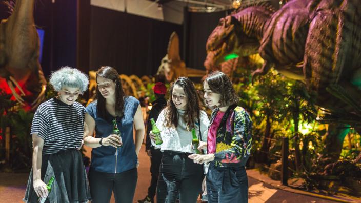 A group of people drinking and standing around an animatronic dinosaur.