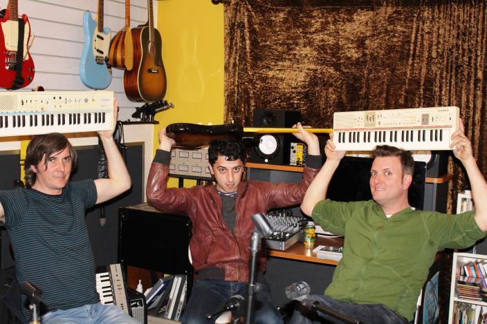 Three men sit in a recording studio holding casio keyboards over their heads.