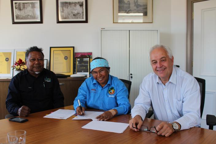 Three people at a boardroom table smile for the camera while one signs a documen