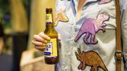 "A close-up of a woman wearing a bedazzled dinosaur shirt. She is holding a beer bottle that incredibly matches her nail polish."