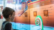 "A young boy looking with awe at a digital screen that has a cartoon of a pirate ship and giant squid on it."
