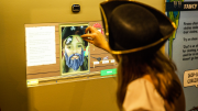 "A girl dressed as a pirate uses a digital interactive that takes a photo of her face, adding illustrated pirate features."