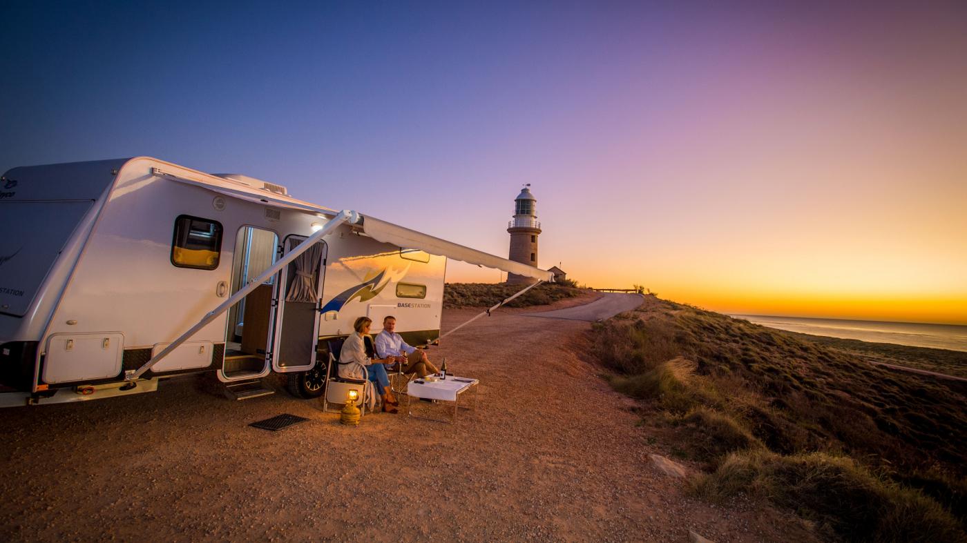 People sit out the front of their caravan with lighthouse behind