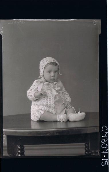 F/L Portrait of baby seated on table wearing cardigan, booties, bonnet; 'Stuber'