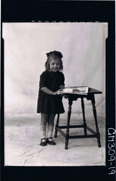 F/L Portrait of girl child standing beside table with book, wearing short dress, ribbon in hair; 'Naggri'