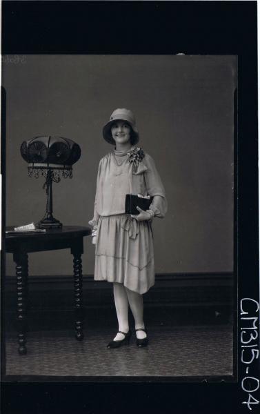 F/L Portrait of woman standing wearing knee length dress, corsage, hat and holding handbag 'Michell'