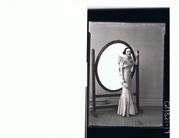 F/L Portrait of woman standing in front of mirror in long evening dress with frills at top & flares at bottom; 'Mills'