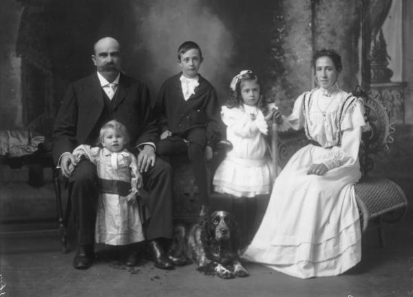 Mrs H Gaston, Group studio portrait of woman wearing long dress seated, girl wearing ribbon in hair standing, boy seated, man seated with child standing and Cocker spaniel dog on floor.