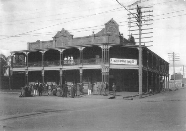 Railway Hotel Kalgoorlie with man and camels being welcomed.  Also shows Banner which reads 'Kalgoorlie Racing Club, Easter Saturday March 30, First Race 2 p.m.' Writing on Hotel facade: 'Wilkies Railway Hotel, AD 1897.' Forrest Street Kalgoorlie opposite Railway Station. 'Sun' Newspaper Office next door.  1919