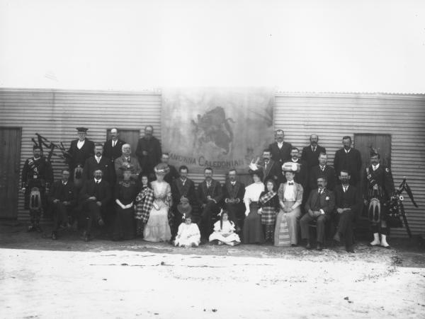 Group of people outside Kanowna Caledonian Society, D.McKellar, banner hanging from roof.