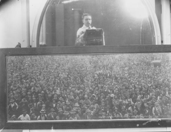 Mackay copying Dwyers famous photo of crowd listening to Father Long announcing locality of Sacred Nugget at Kanowna.  Copy, note photographer Mackay taking photo in mirror.