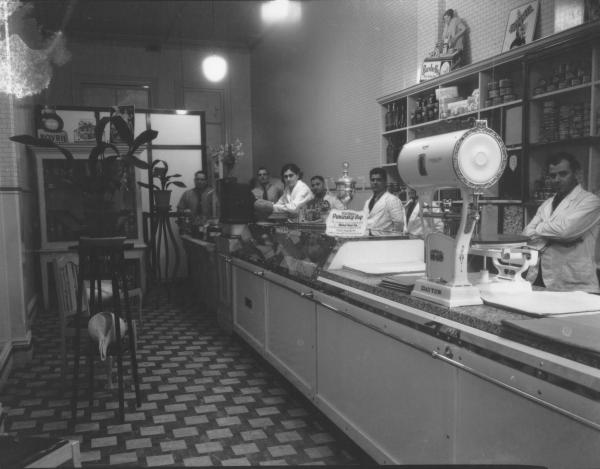 Interior of Kalaf's shop with footballPremiers Cup on counter donated by George Calaf.  Large CAYTON scales on counter, 'Bushells Tea' ad. on display with various other products in tins and jars. 1 woman and 5 men behind counter.