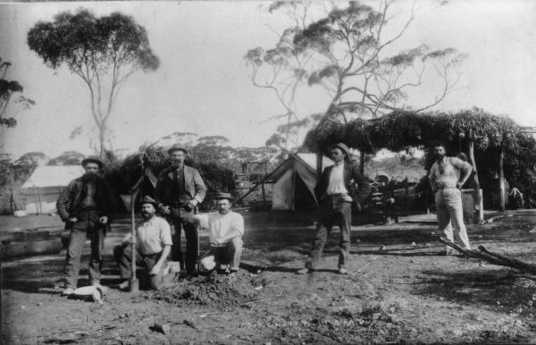 Early days mining camp. (for Mr. Reid.)  Alf Kyle's lease, 25 mile. Doncaster Camp.  C Mason, Col Cameron,kneeling,  standing L to R -  E Thheile, C Brown, Jerome de Jaries, Alf Kyle.  Tents and bush shelters in background.