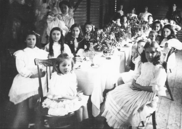 Children seated on house verandah(?) at a long table decorated for a party. Party dresses - frilled lace etc. VICKERS