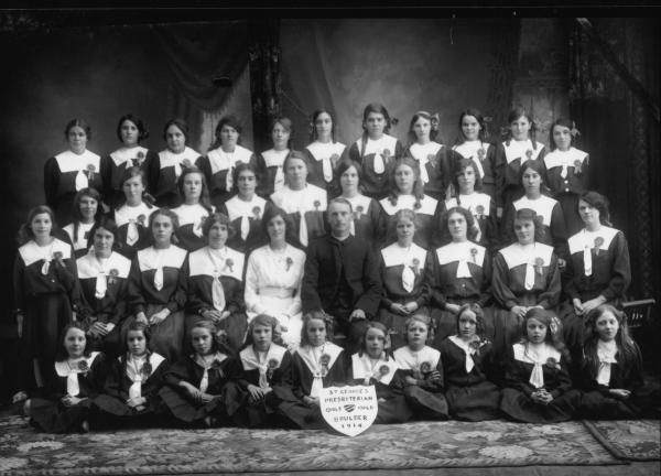 Studio group photograph of clergyman and girls wearing dark dresses with white sailor collars and rosettes. Shield inscribed 'St George's presbyterian Girls Guild Boulder 1914.