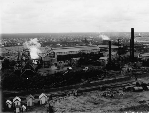 View of minesite with Boulder City in background. Wood stack and rail track in foreground. Miners' canvas / hessian houses also in left foreground.