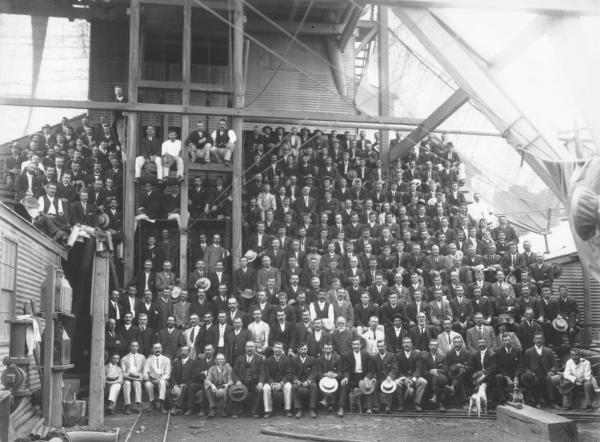 Large group of miners and staff at Kalgurli Goldmine sitting under headframe, most have taken off hats and are holding them.Frederck George Brinsden (Born 1980 in Creswick Vic, died 1958) is second on left in front row.