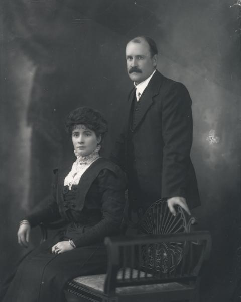 3/4 portrait of woman seated and man standing, both wearing formal black clothes, woman with jewellry. 'Mr. A.E. Sharland'
