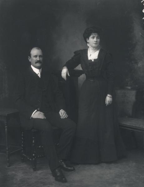 F/L portrait of man seated and woman standing, both wearing formal black clothes, woman with jewellry. 'Mr. A.E. Sharland'