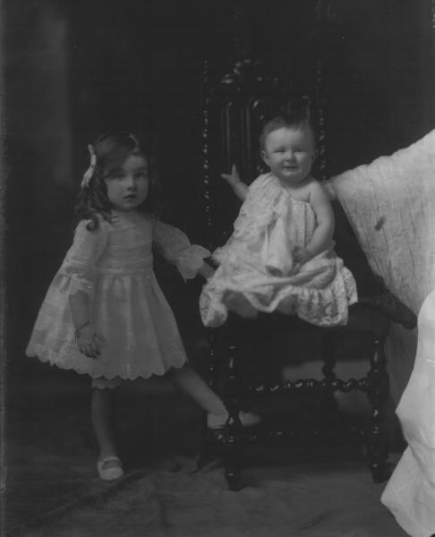 F/L portrait of 2 young children. Girl standing  wearing short dress, baby seated wearing long lace gown. Mrs. C. Reeves.