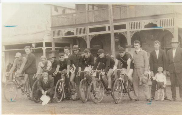 Menzies to Kalgoorlie Bike race taken outside Grand Hotel, Menzies. 6 men on racing bikes with several other men and one child.  Montgomery's store in the background.