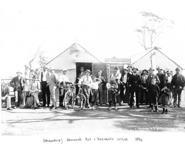 Group of men and children outside 'Post Telegraph Office'Two men have push bikes. Building made from hessian and wood.