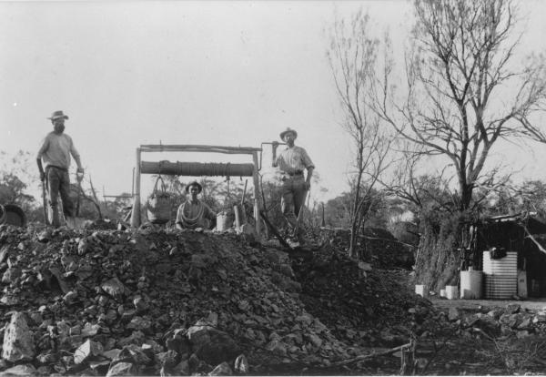 Three men standing on mine workings with hand winder, bucket and various tools, low store shed by shaft and rain  water tank.