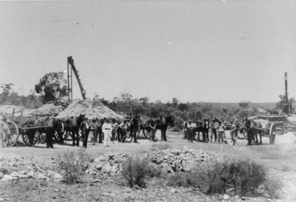 Men with horses and carts posing by 2 small mines, mackine 'poppet heads', slag heaps. Most men in working clothes with shovels, one man in suit and hat, another in white pants and coat and bandana type belt posing in centre of line.