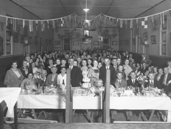 Large group of men, women children standing in hall by table set for wedding feast, 2 tiered celebration cake and other cakes on table with crockery, cutlery, drinks, flowers.Banners strung diagonally across room.