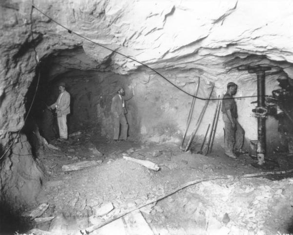 Mine, underground, 3 men, mining tools propped by wall, hoses, air pipes, candles.