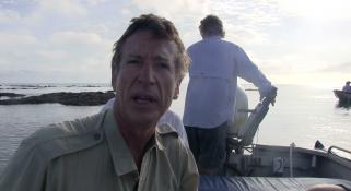 Clay Bryce talking to camera sitting in a small boat with man standing behind him steering