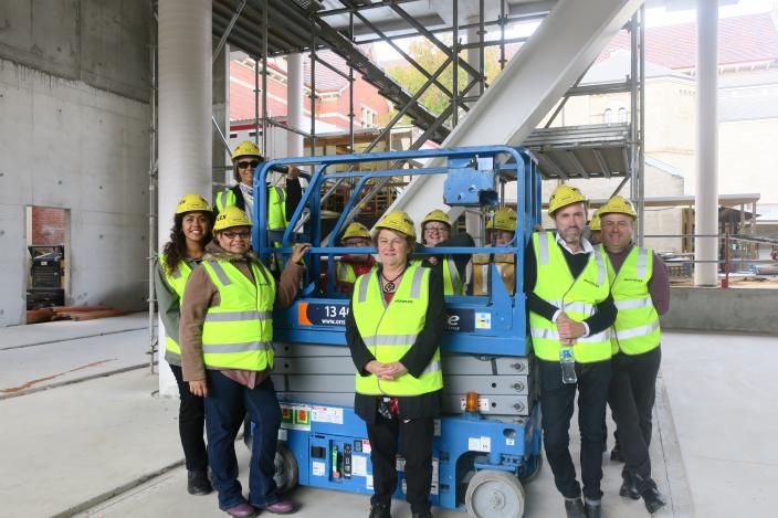 "People in yellow high visibility jackets stand around a blue scissor lift."