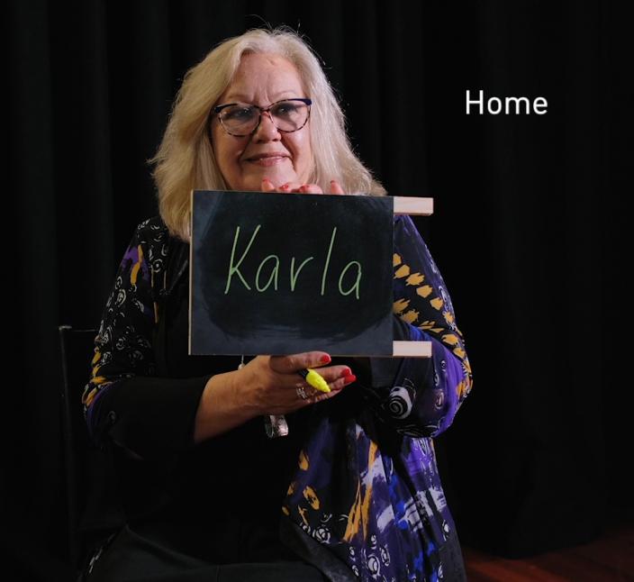 "A woman holds up a small chalkboard that reads 'Karla'. Text reads 'Home'."