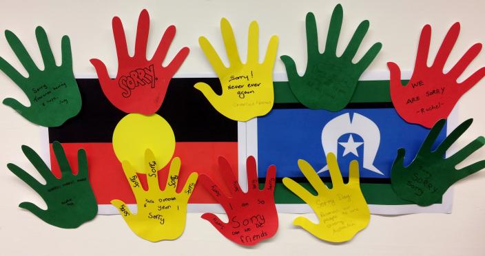 "10 hands are cut out from coloured paper with apologies written on them. They are stuck around Aboriginal and Torres Strait Islander flags."