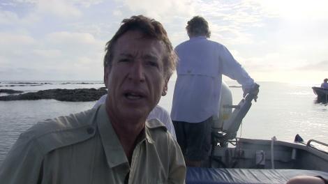 Clay Bryce talking to camera sitting in a small boat with man standing behind him steering