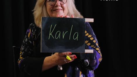 "A woman holds up a small chalkboard that reads 'Karla'. Text reads 'Home'."