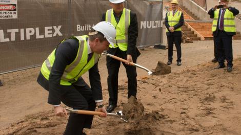 Premier McGowan bending with a ceremonial shovel and Minister Templeman standing to his left.  They are turning sod by digging the soil at the site of the New Museum.  Both are wearing fluro yellow vests and hard hats.