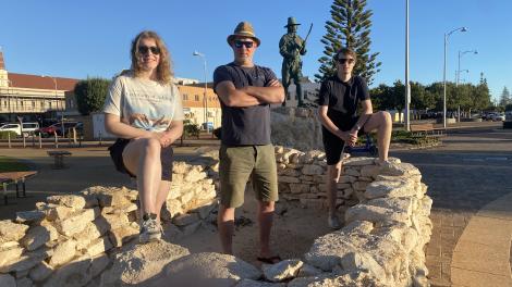 Three people stand on a circular stone structure with a statue and a tree in the background.
