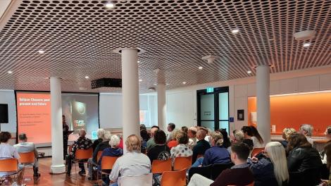 A large group of people seated in orange and white chairs face a large projector at the front of the room with an individual standing at a podium. The image projected onto the screen displays a book titled 'Eclipse Chasers' and some smaller text