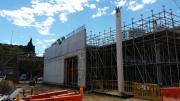 A concrete wall stands at the background of the photo. A pole is in the foreground.  Scaffold is holding up the structure. 