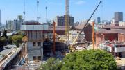 "Tree in foreground, concrete square block around three storeys high, cranes and machinery moving and lifting concrete."