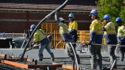 Six workers in high vis and hard hats stand waiting for concrete to pour.  Two workers holder a hose and pour the concrete into position on the ground. 