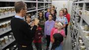 "A group of children stand listening to a man holding a jar, they stand amongst metal shelves of jars."