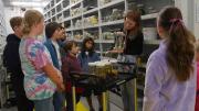 "A woman stands holding a jar with a reptile in it while children stand listening to her, a trolley holding more jars is in front of her.