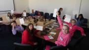 "A group of children sit around a large table, smiling and showing pages of their notebooks to the camera."