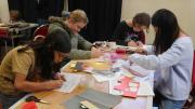 "A group of children sit and stand around a trestle table, drawing and cutting pieces of paper."