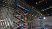 Two scissor lifts fully expanded have men atop working on the ceiling of a large room.