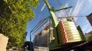 A yellow crane lifts a large section of steel over a heritage building. A tree is next to the crane, the sky is blue